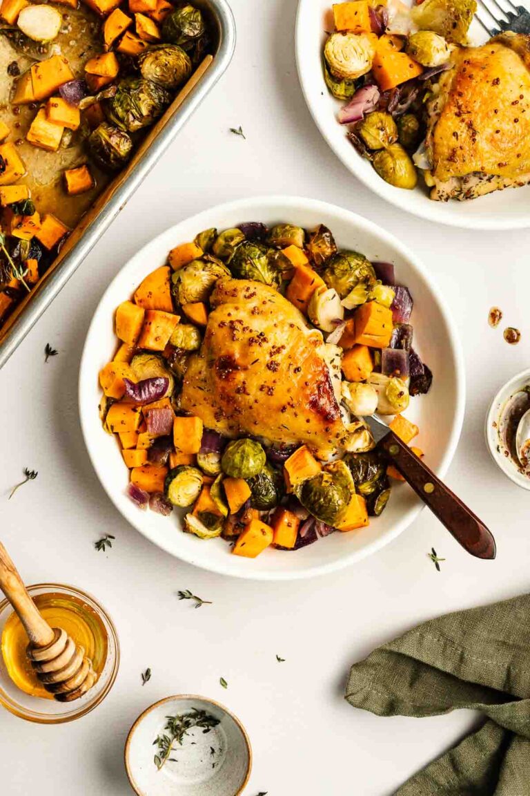 A plate of roasted chicken thigh served with Brussels sprouts, sweet potatoes, and red onions. A sheet pan with more roasted vegetables and a bowl of honey are visible nearby.