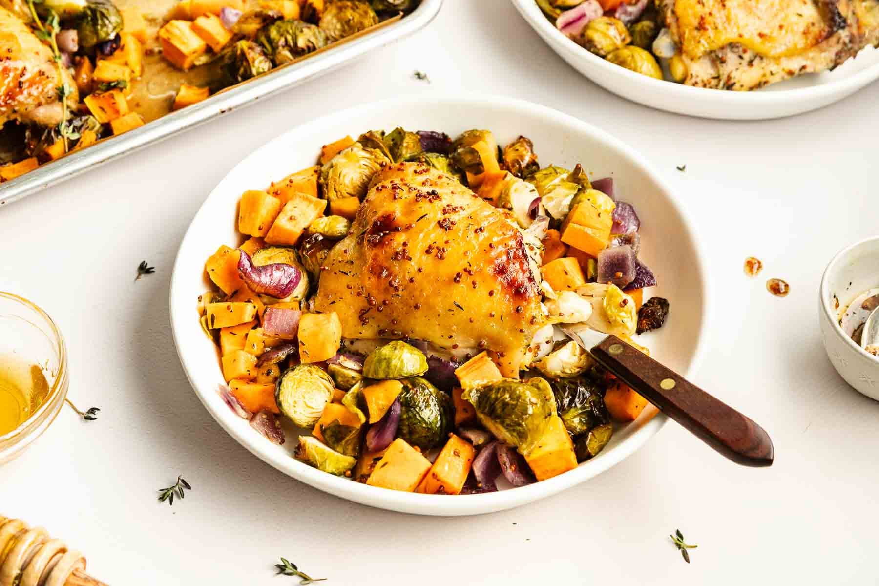 A white bowl holds roasted chicken thigh, Brussels sprouts, sweet potatoes, and red onions, with a knife on the side. Other servings and a small sauce bowl are nearby on a white table.