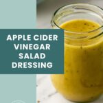 A glass jar filled with yellow apple cider vinegar salad dressing sits on a marble surface next to a blue-green label with white text.
