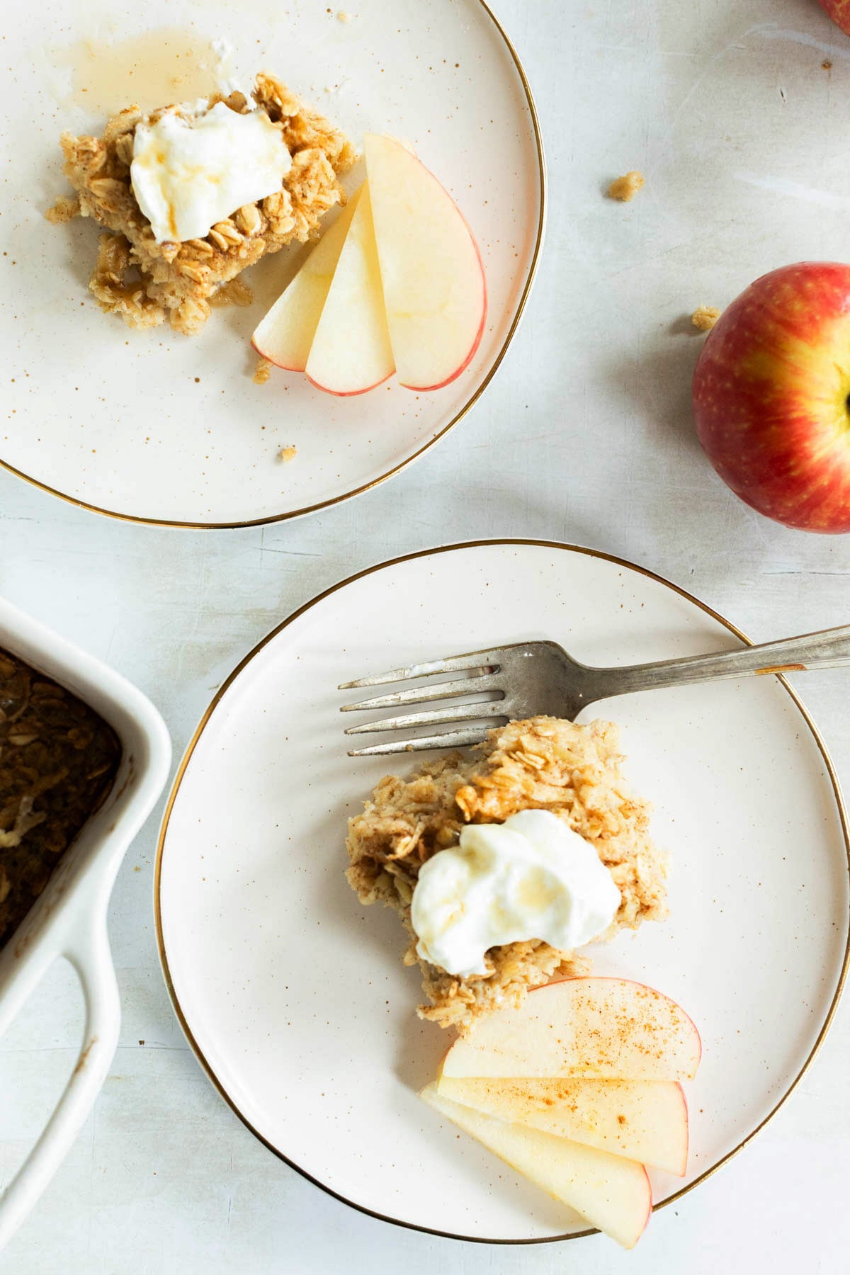 Two plates with baked oatmeal topped with yogurt and apple slices, a fork, a whole apple, and a baking dish are on a white surface.