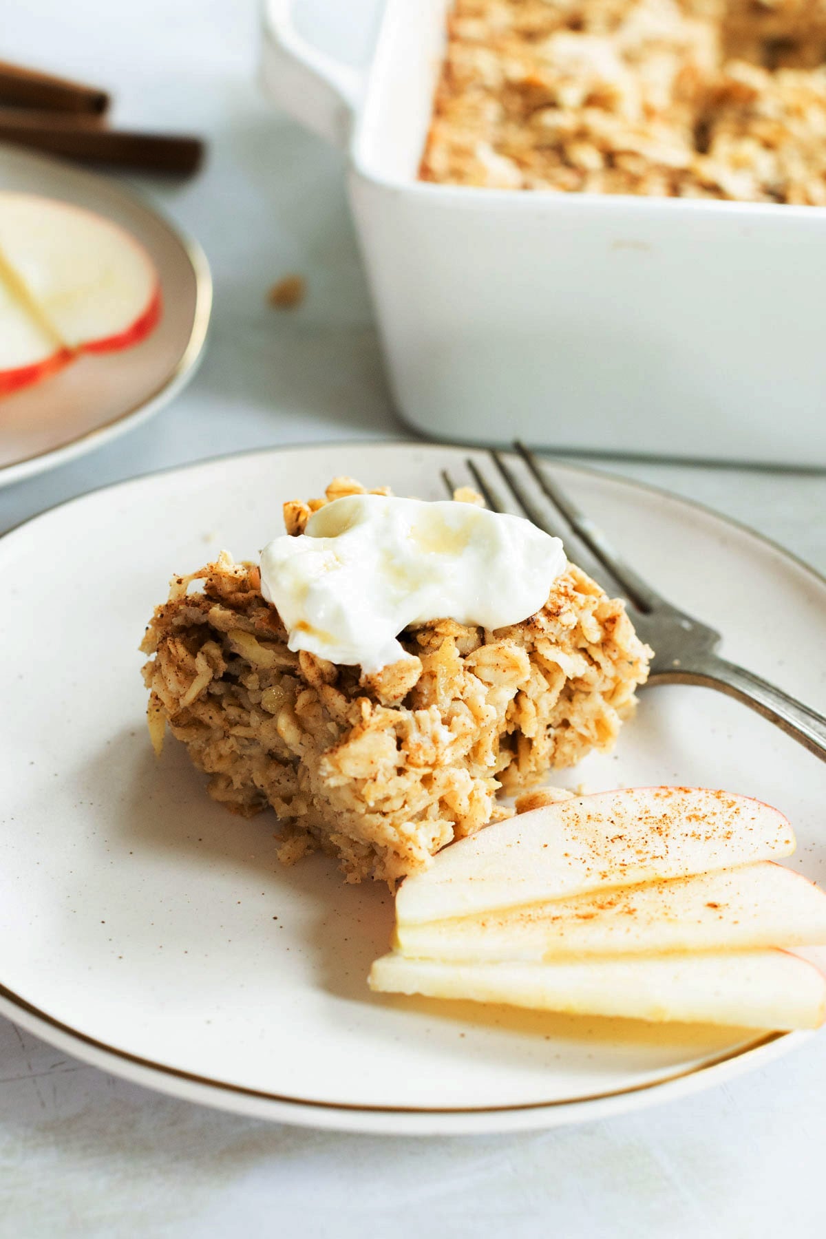 A square serving of baked apple oatmeal with a dollop of yogurt and apple slices on a white plate, with a fork and a casserole dish in the background.