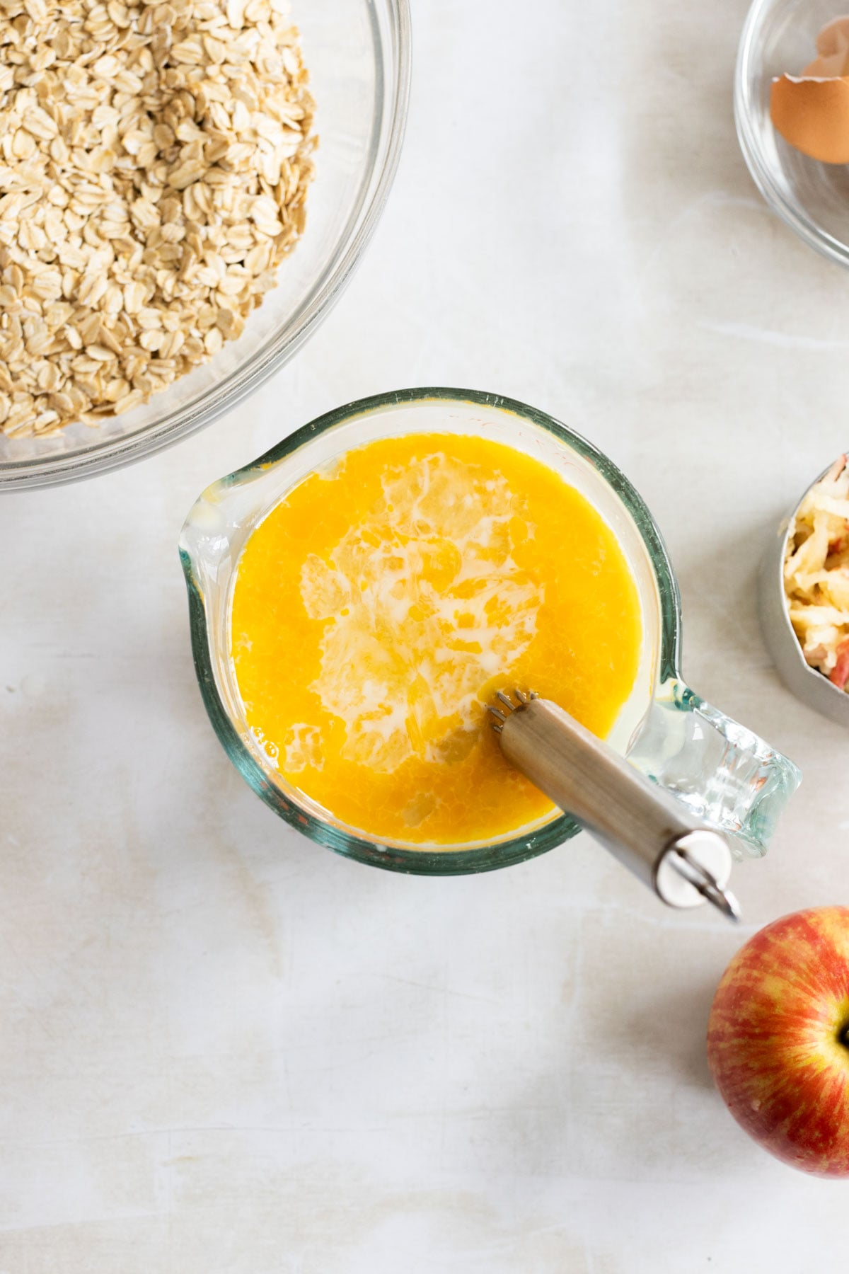 A glass measuring cup with a yellow liquid mixture and a whisk, surrounded by a bowl of oats, an apple, and a cracked egg.