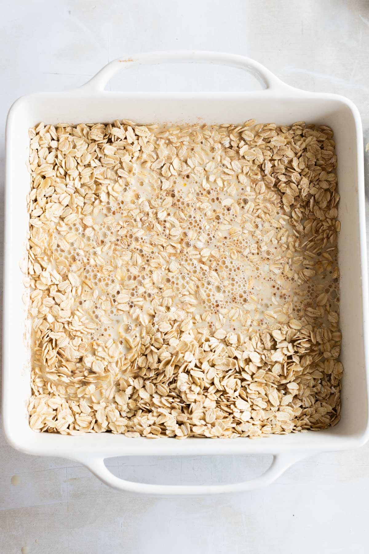 Square white baking dish filled with uncooked rolled oats and milk mixture, sitting on a light-colored surface.
