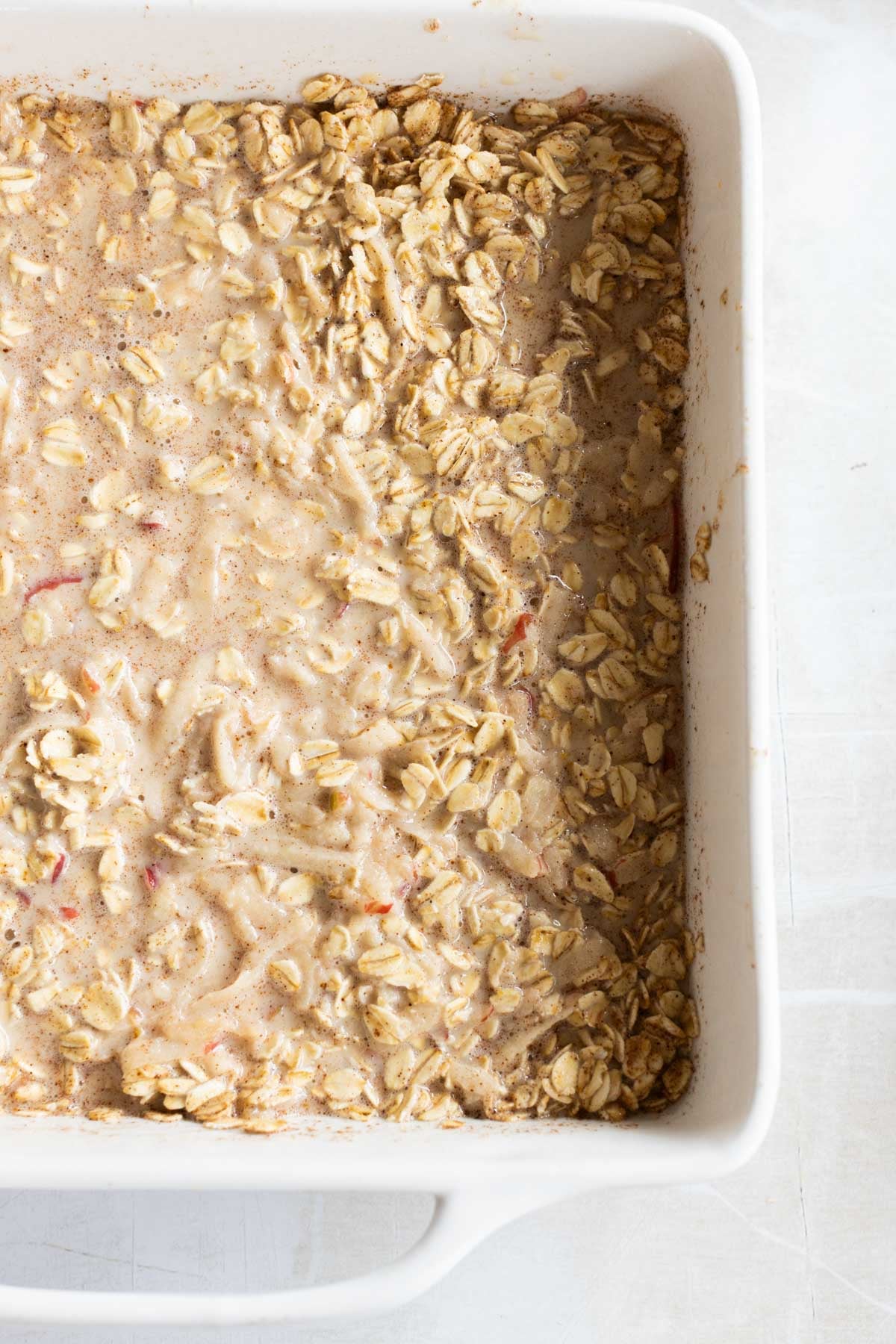 A white baking dish filled with unbaked apple oatmeal mixture, featuring visible oats and liquid, sits on a light-colored surface.