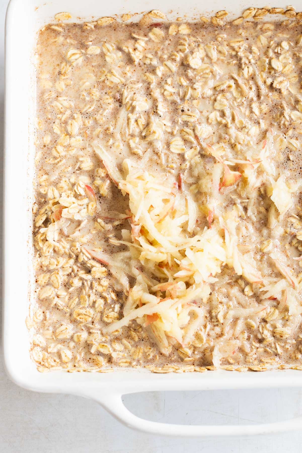 A white baking dish filled with an oatmeal mixture topped with a pile of shredded apples, ready to be baked.