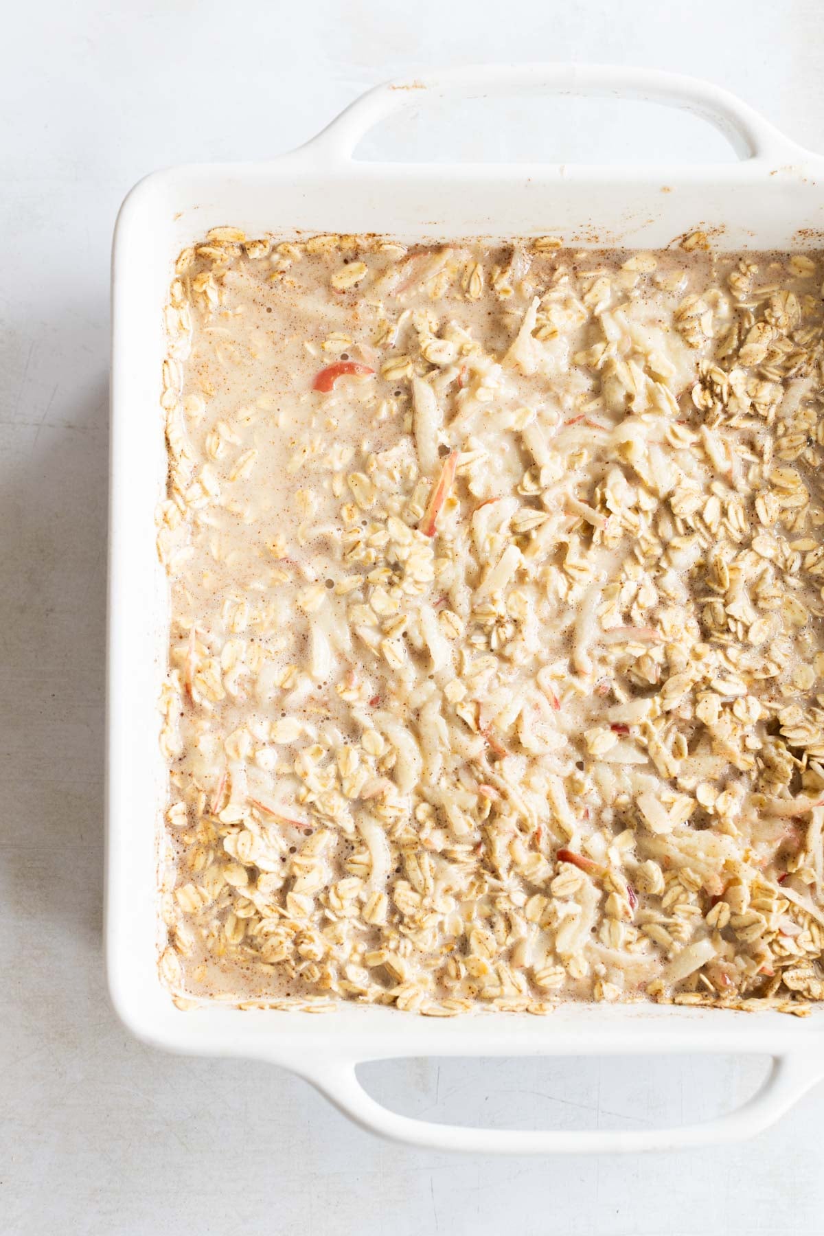 A white baking dish filled with baked oatmeal containing oats and visible pieces of apple, sitting on a light surface.