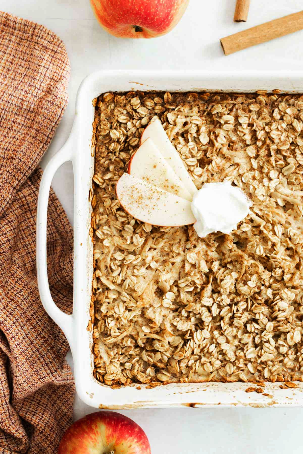 Baked apple oatmeal in a white baking dish, topped with apple slices and whipped cream. Nearby are apples, a cinnamon stick, and a textured cloth.