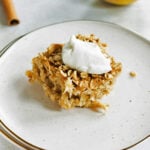 A square serving of baked oatmeal topped with a dollop of yogurt on a white plate, with apples and a cinnamon stick in the background.
