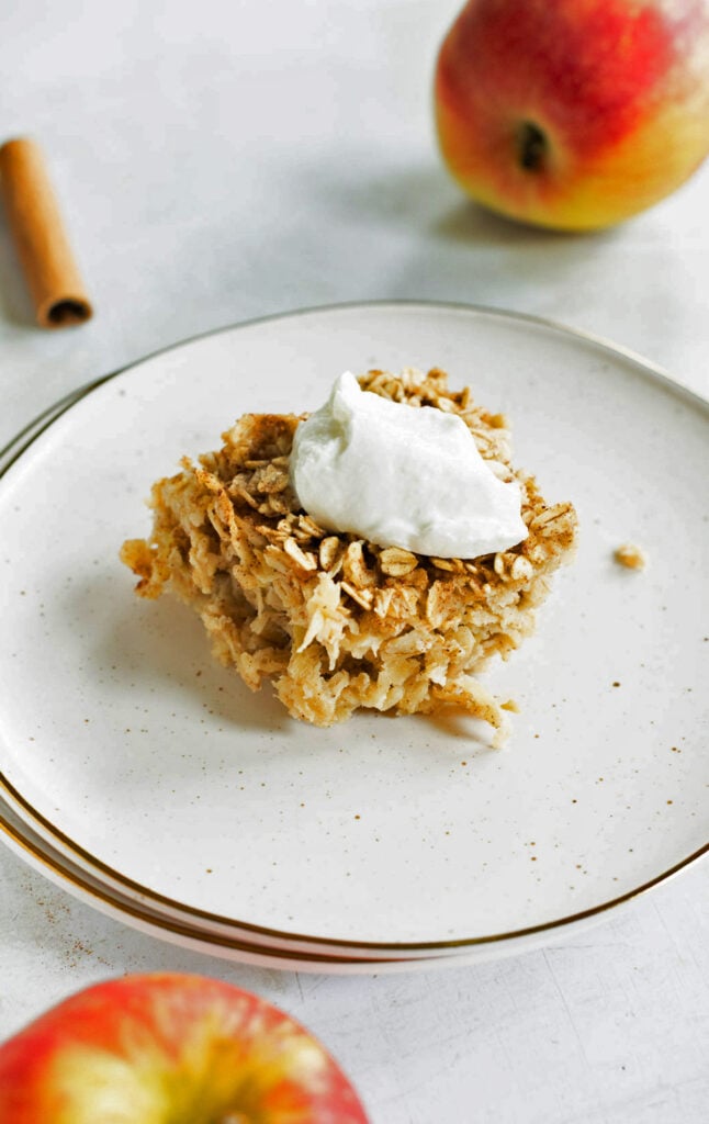 A square serving of baked oatmeal topped with a dollop of yogurt on a white plate, with apples and a cinnamon stick in the background.