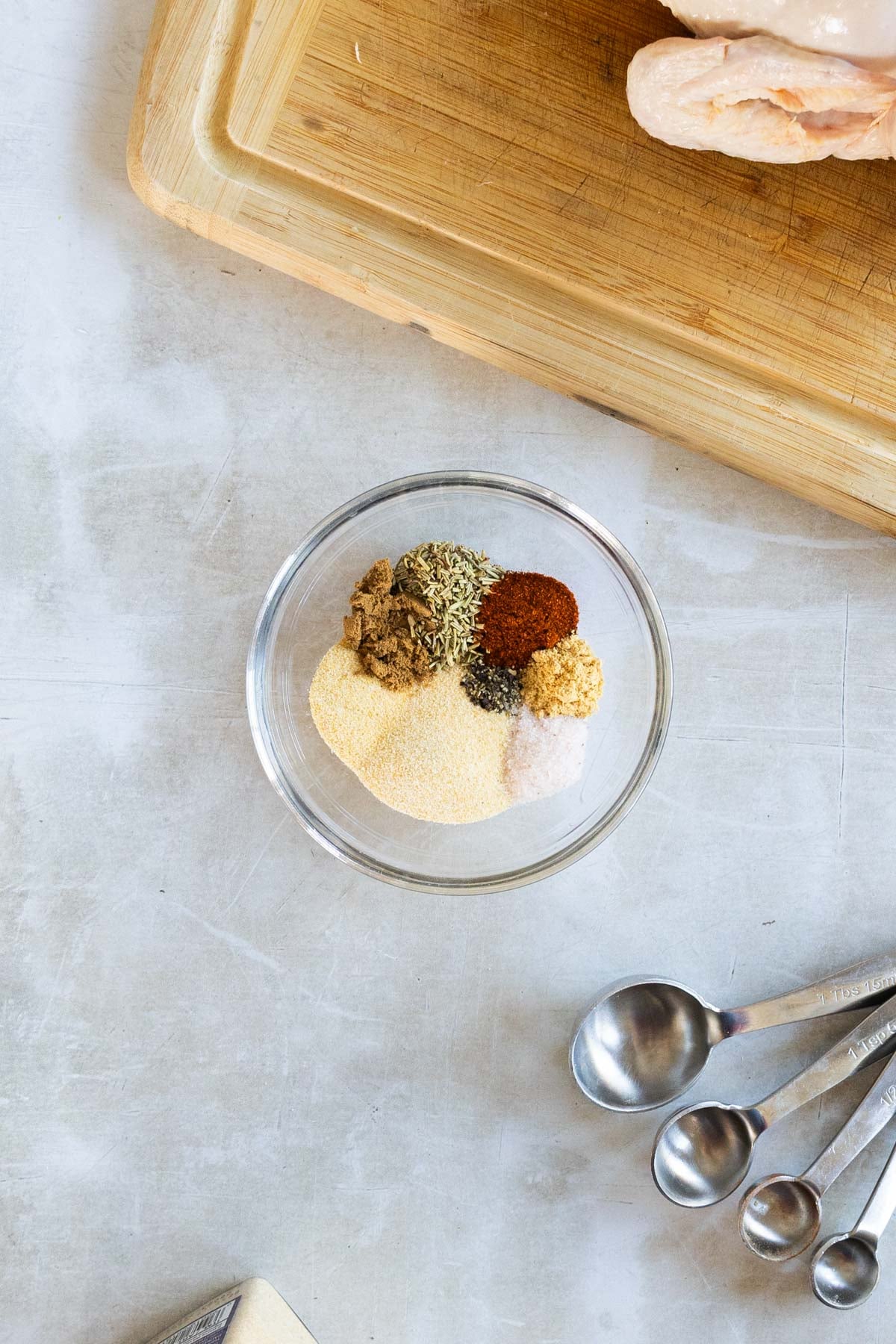 A glass bowl with assorted spices and seasonings next to metal measuring spoons on a light countertop, with a wooden cutting board partially visible at the top.