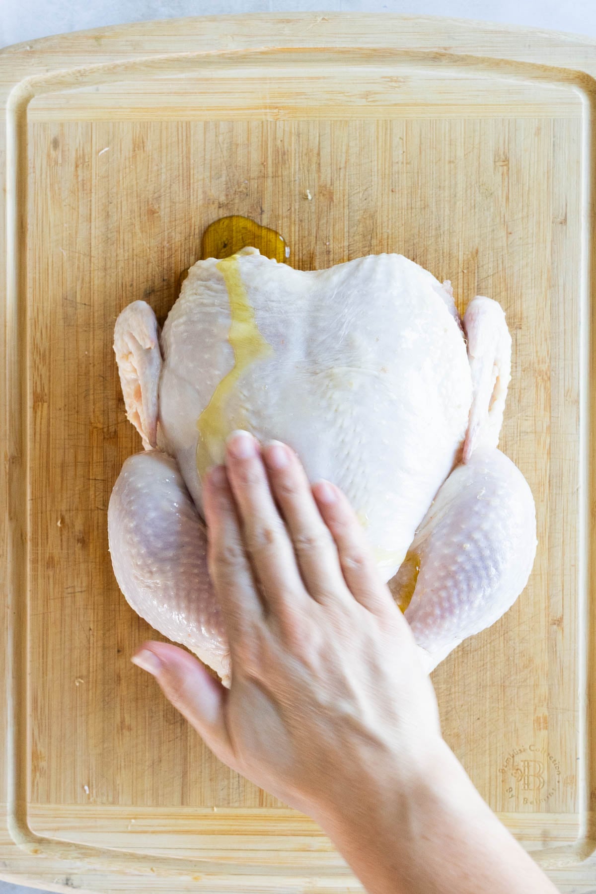 A hand rubs oil onto a raw whole chicken placed on a wooden cutting board.