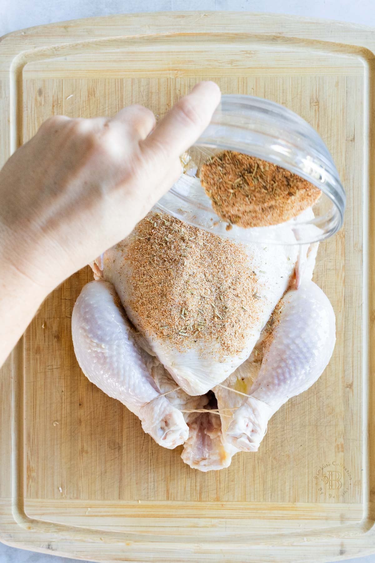A hand sprinkles seasoning onto a whole raw chicken placed on a wooden cutting board.