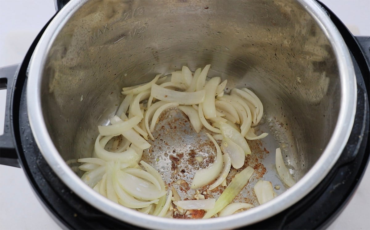 Sliced onions being sautéed in a pressure cooker, with browned bits stuck to the bottom of the stainless steel inner pot.