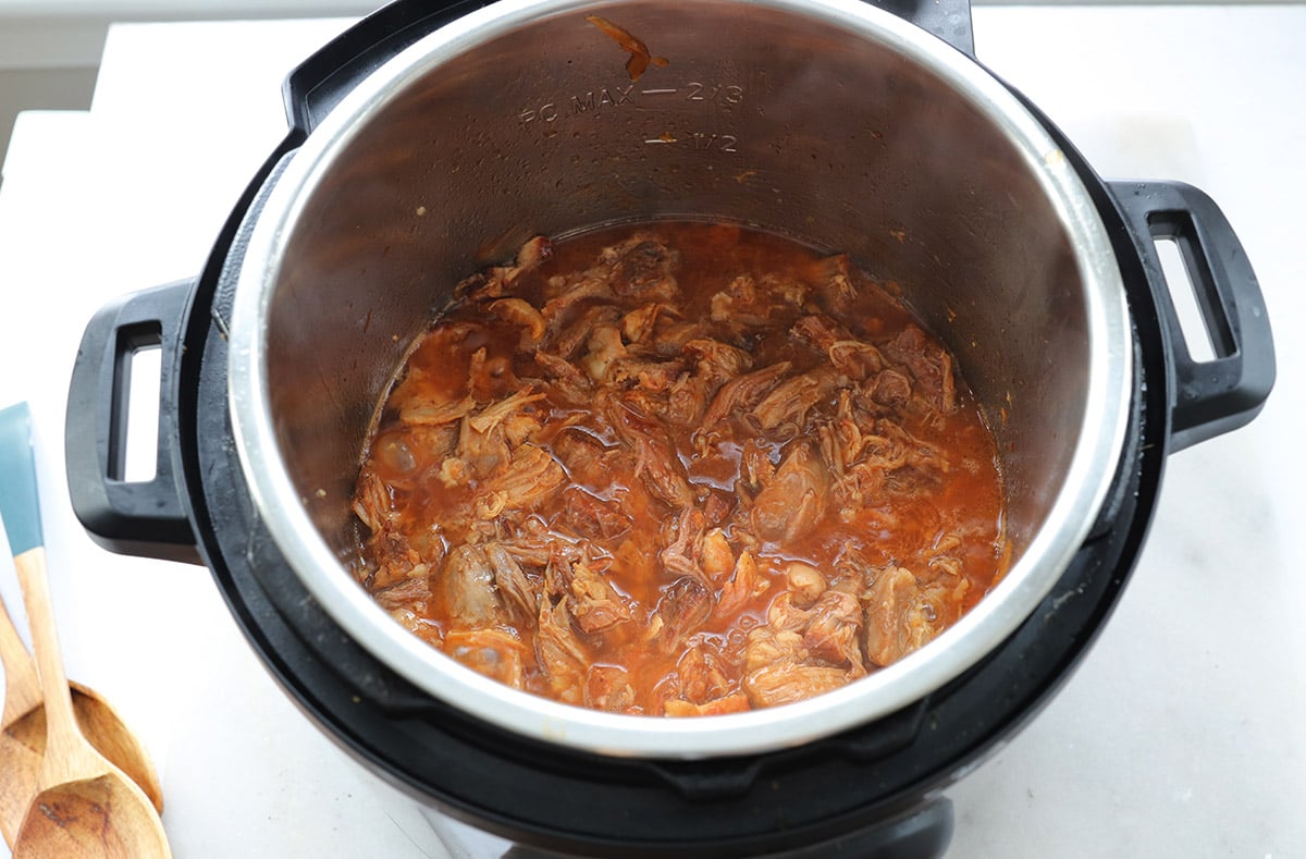 Shredded meat in red sauce inside an open pressure cooker, with wooden utensils on the counter nearby.