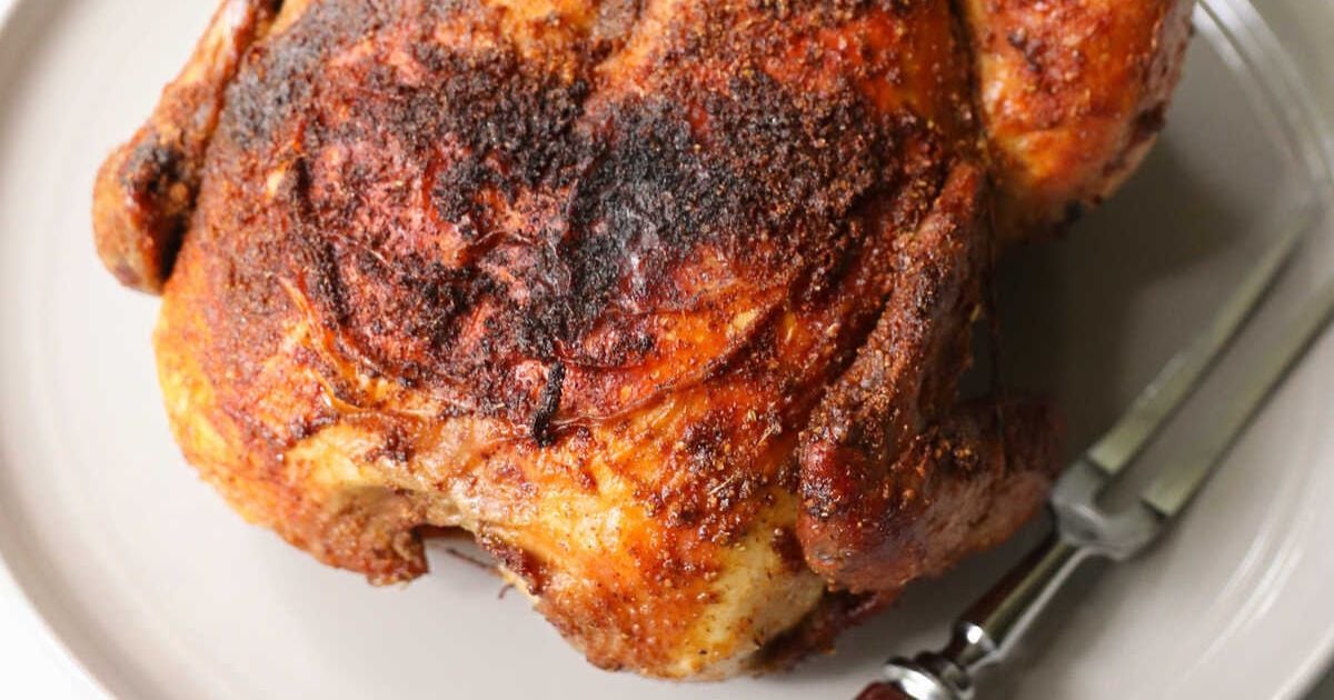 A whole roasted chicken with crispy, seasoned skin sits on a plate next to a serving fork.