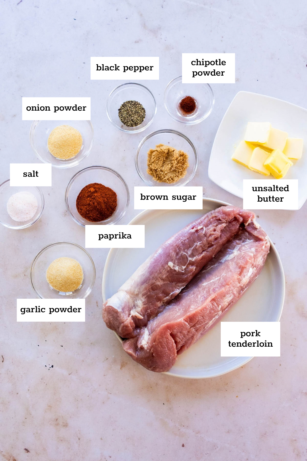 Raw pork tenderloin on a plate surrounded by bowls of salt, onion powder, garlic powder, paprika, black pepper, chipotle powder, brown sugar, and butter.