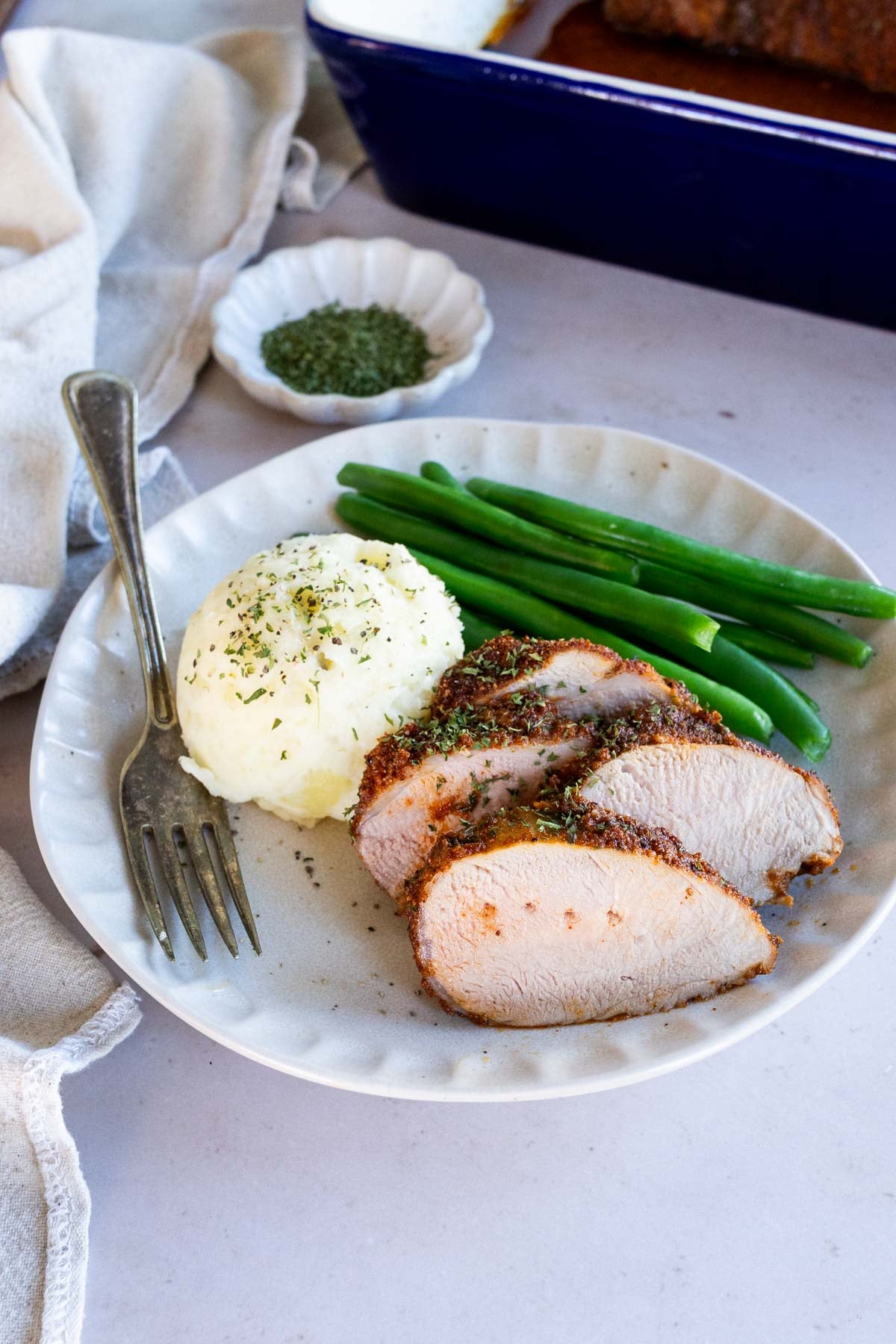 Slices of baked boneless pork with mashed potatoes and green beans on a plate, with a fork and a small dish of herbs nearby.