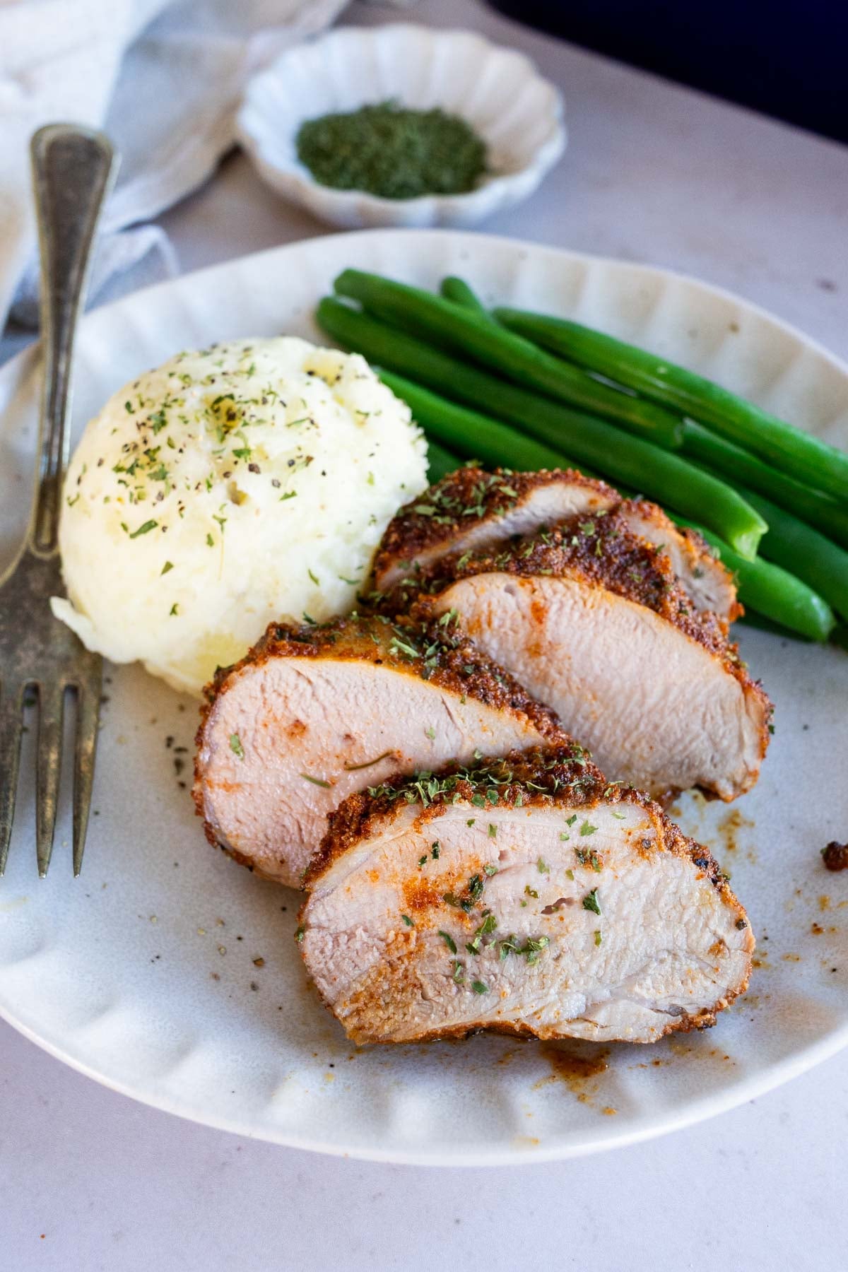 Sliced pork tenderloin with mashed potatoes and green beans on a white plate.