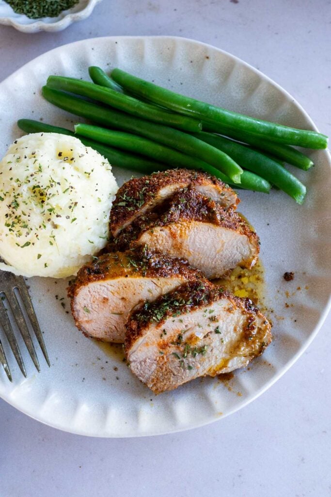 Sliced seasoned baked pork tenderloin served with mashed potatoes and green beans on a plate, viewed from overhead.