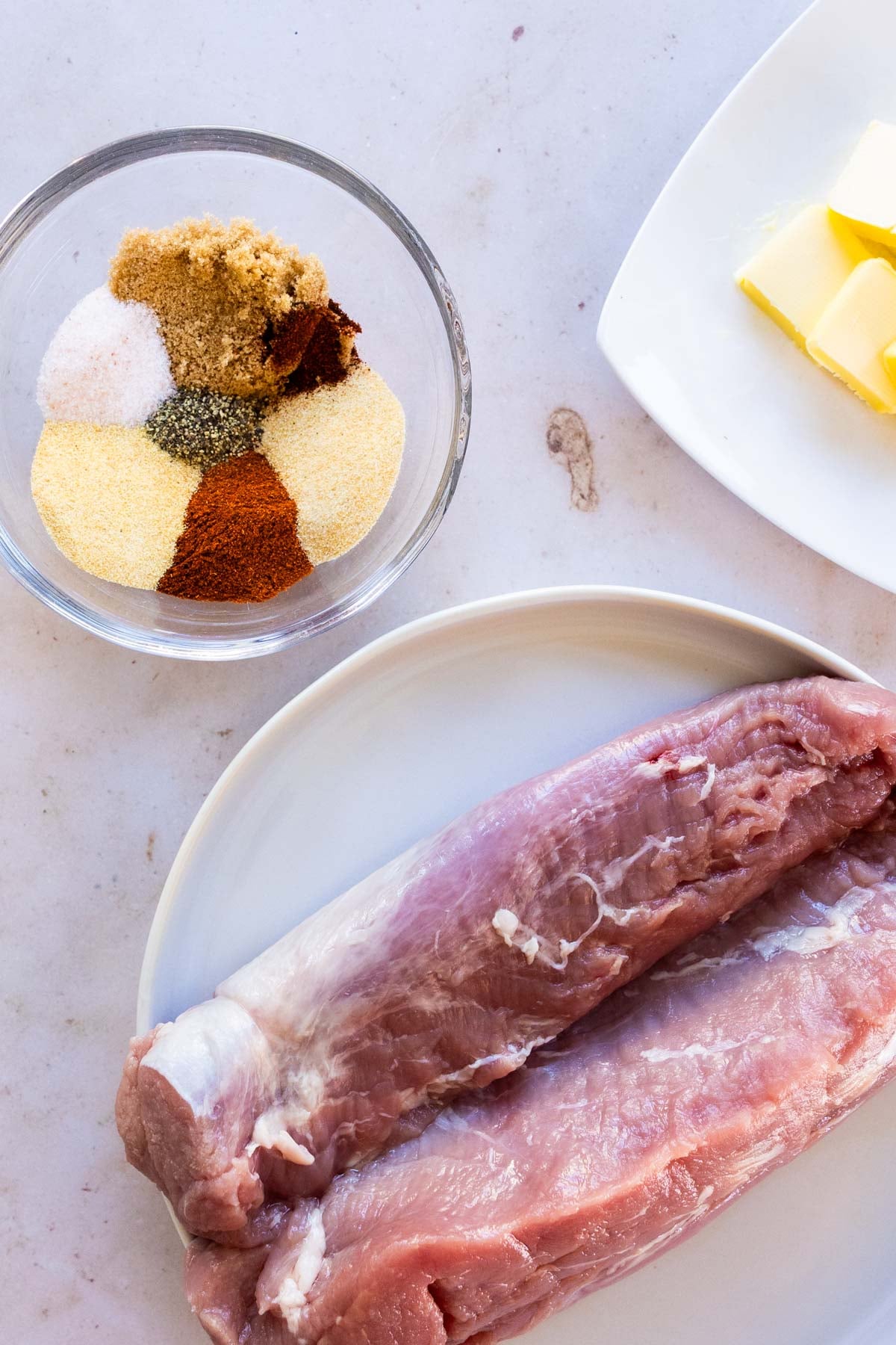 A raw pork tenderloin, a glass bowl containing various spices and brown sugar, and pieces of butter on a white surface.