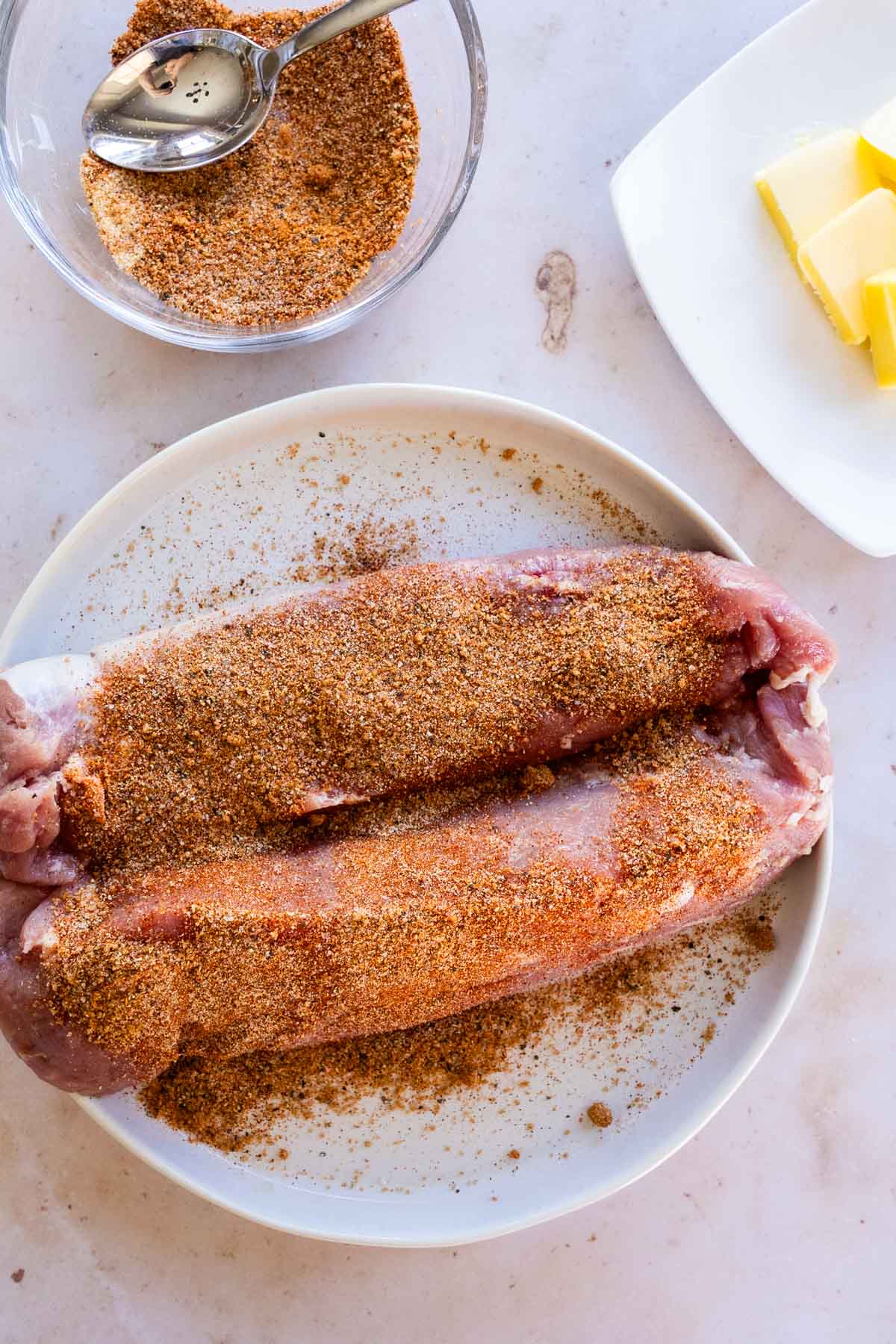 A raw pork tenderloin on a plate, coated in dry spice rub, with a bowl of the seasoning and butter slices nearby.