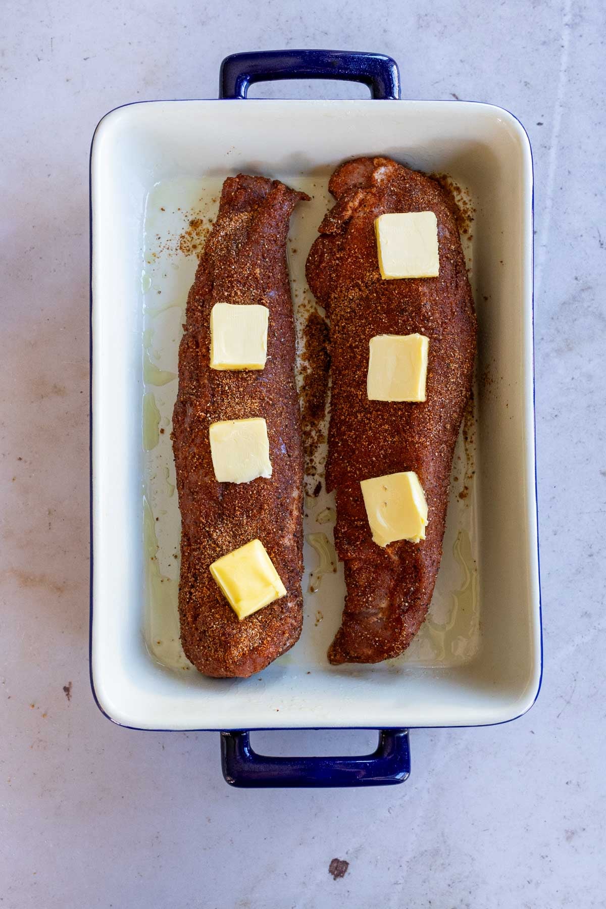 Two seasoned pork tenderloins topped with pats of butter in a ceramic baking dish.