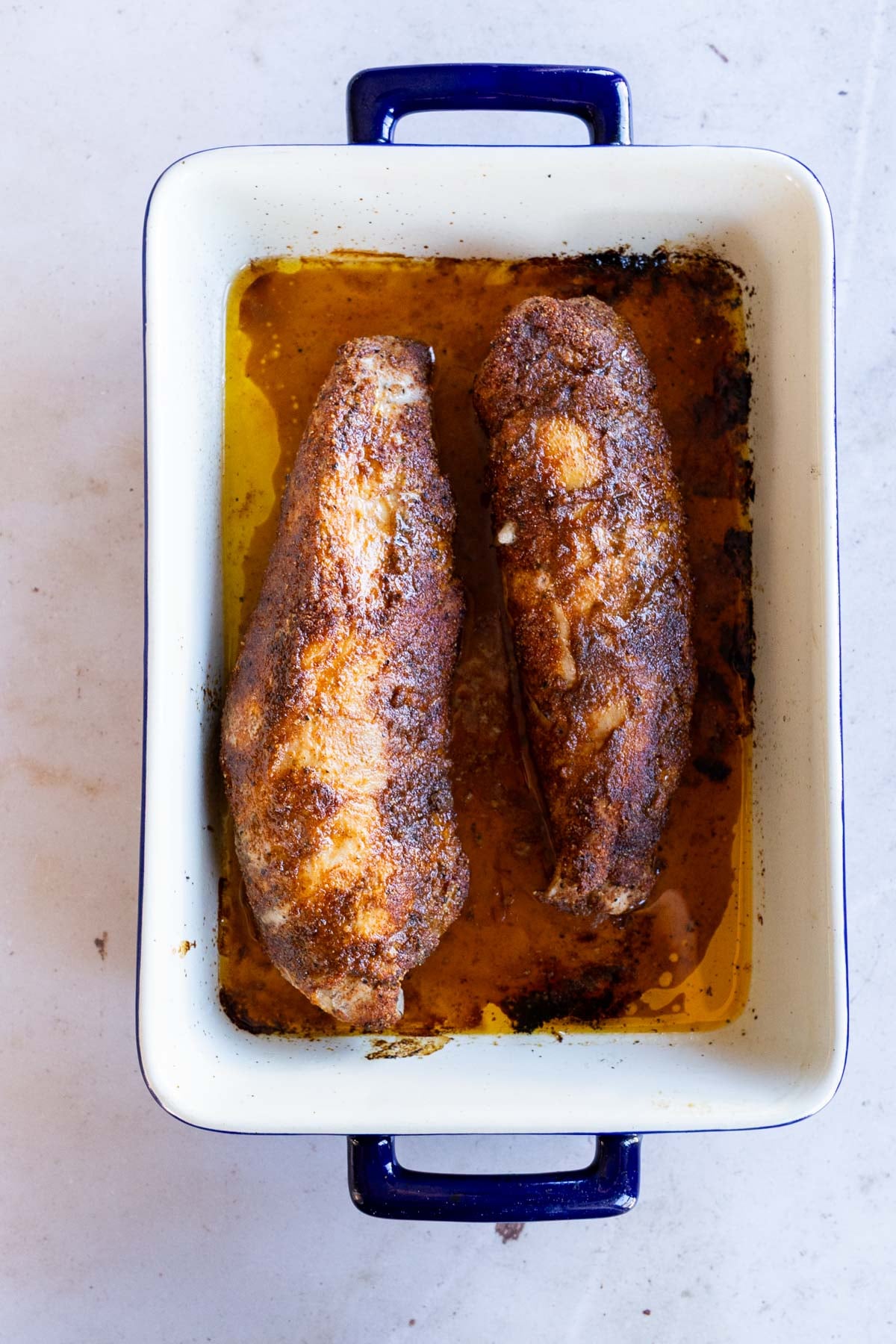 Two seasoned pork tenderloins in a baking dish with pan juices.