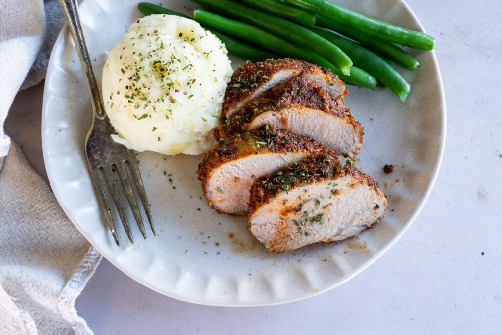 Sliced seasoned chicken breast, a scoop of mashed potatoes, and green beans on a plate with a fork.
